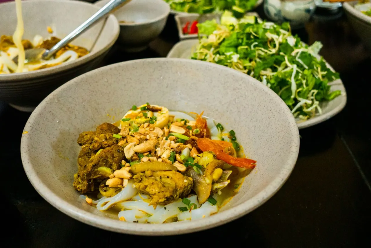 Tô mì Quảng đầy đủ topping, sợi mì vàng nghệ, rau sống ăn kèm bên cạnh