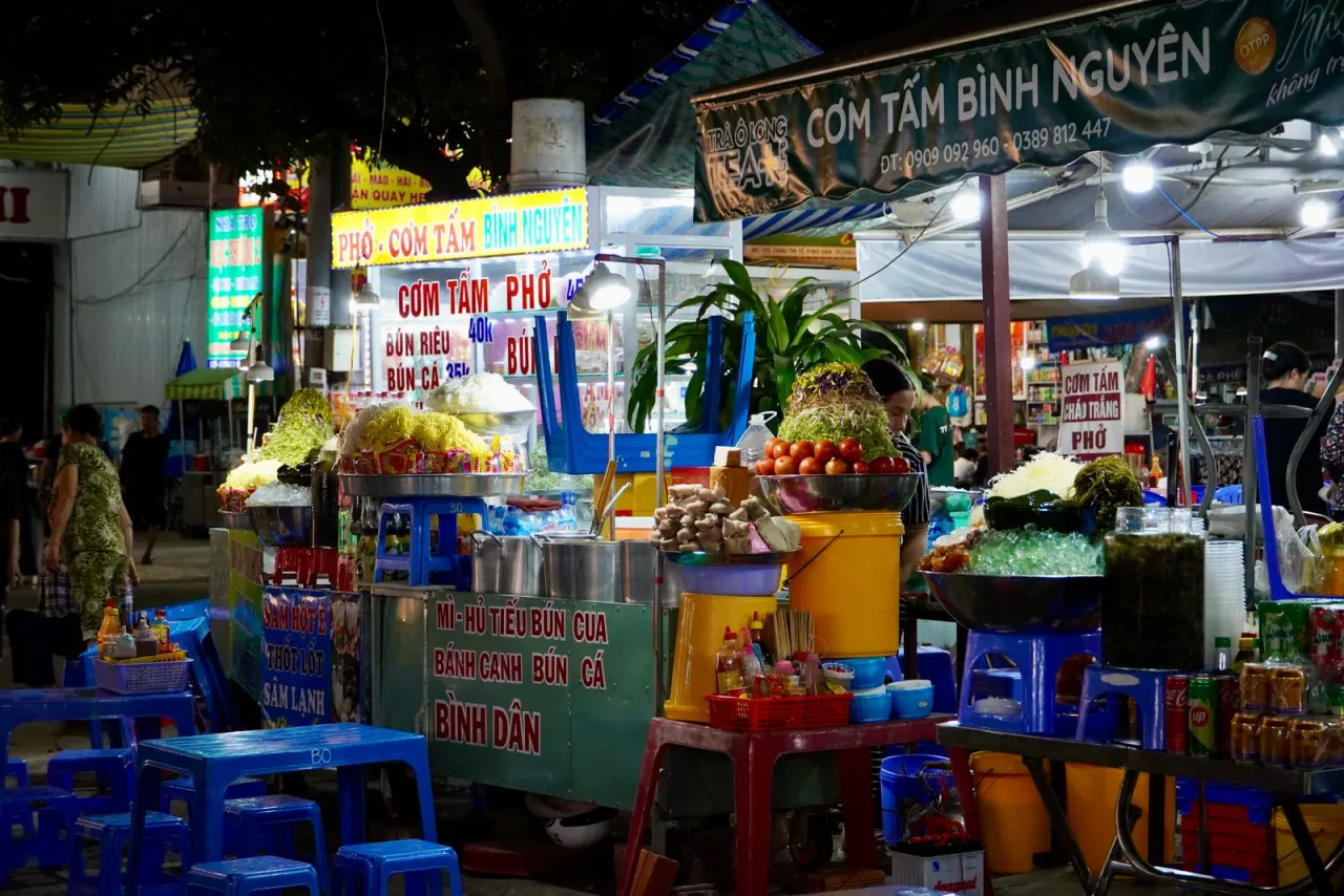 Quầy ăn vặt đêm rực rỡ ánh đèn, bảng hiệu cơm tấm phở, ghế nhựa xanh đặc trưng vỉa hè Việt Nam